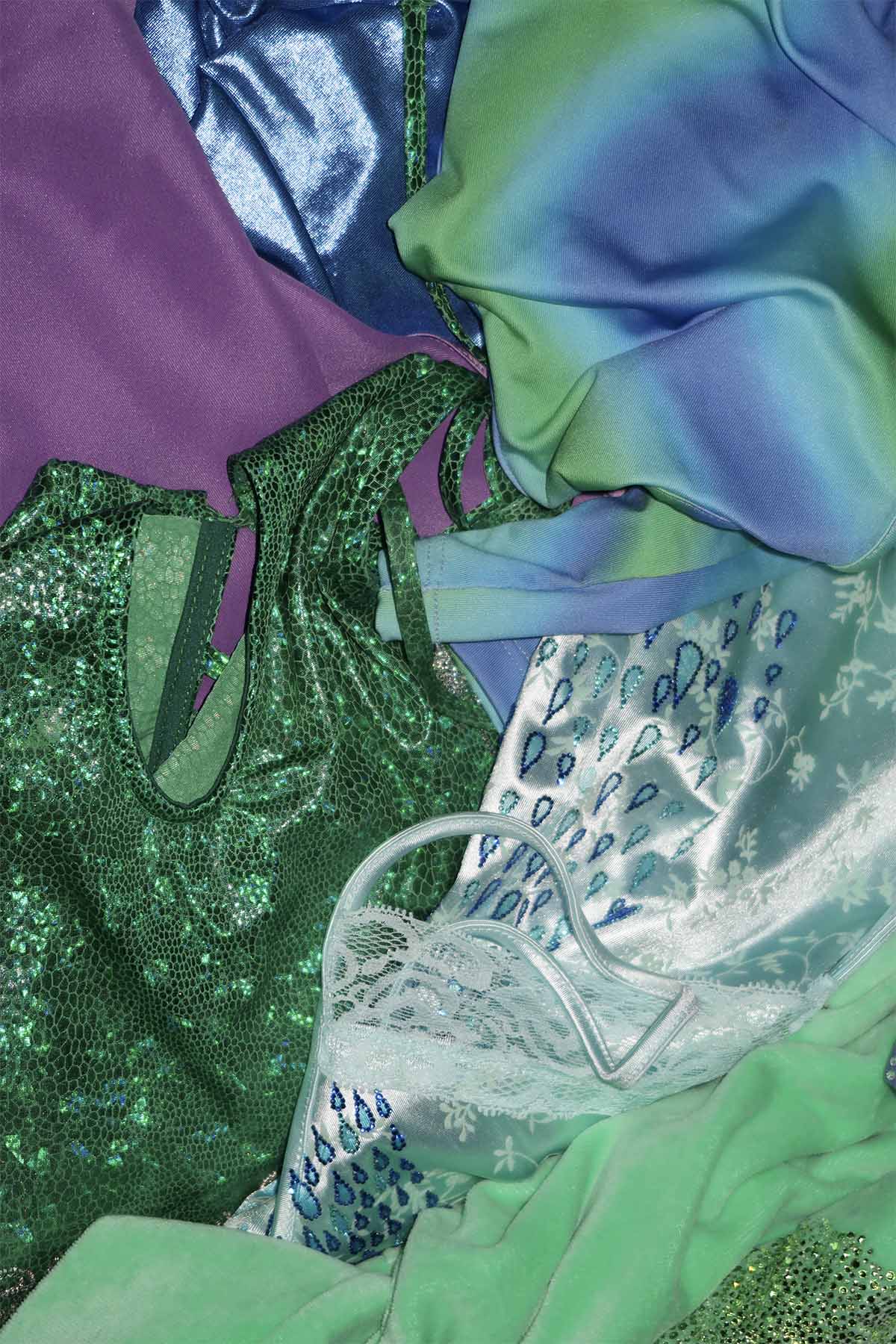 A pile of clothing in bright and metallic shades of blue, purple and green. The pile includes a pale blue satin dress with light blue and dark blue rhinestoned droplet shapes.
