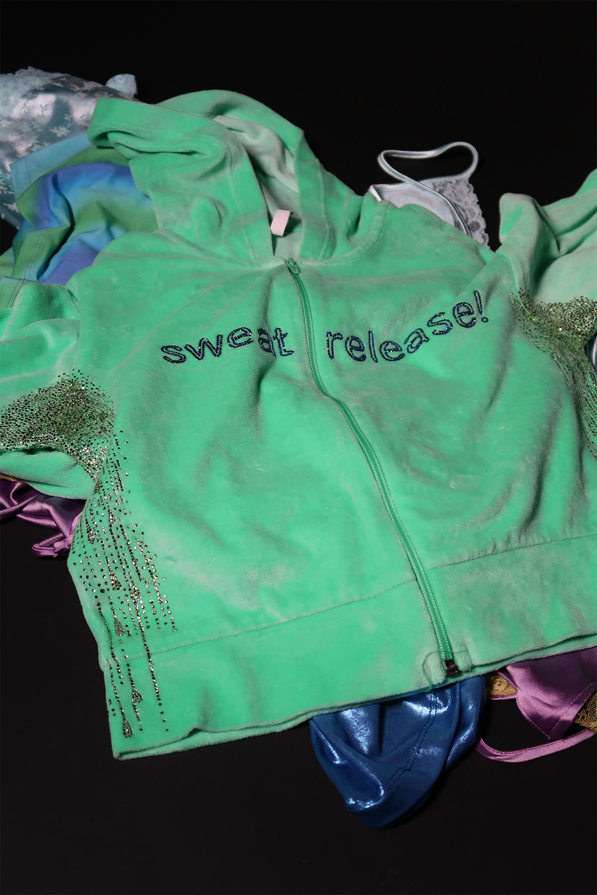 A green velour tracksuit top is at the top of a pile of unworn clothing on a black background. It has the slogan ‘sweat release’ added to the front of the top in dark blue rhinestones. The armpits are covered in rhinestones that mimic sweat patches. These patches trail into droplets as the rhinestones move down the torso of the top.