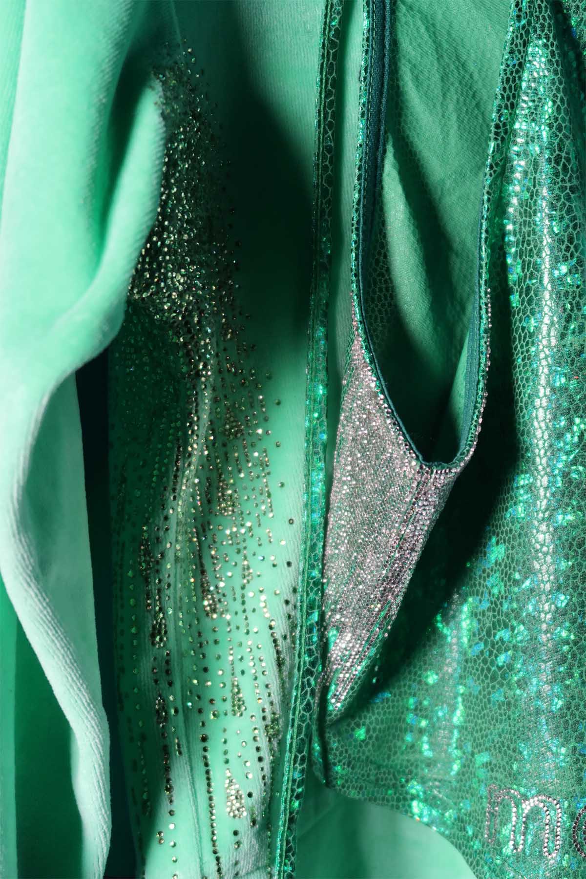 Arm details of an unworn light green velour tracksuit top, and a darker green metallic halter-neck top. The velour top has light green rhinestones attached from the armpit in droplet shapes. The metallic top has silver rhinestones attached from the underarm in straight lines.