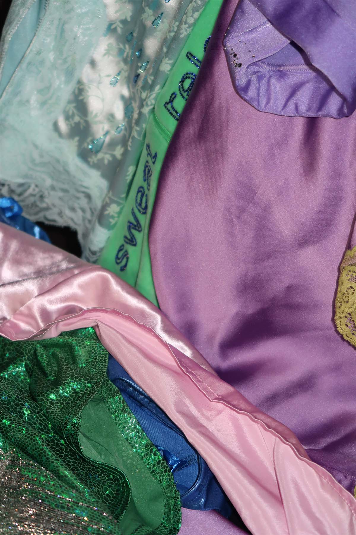 A range of blue, pink, purple and green clothing layered over each other. Some of the garments have rhinestones added, and glimpses of these are visible within the layers of clothing.