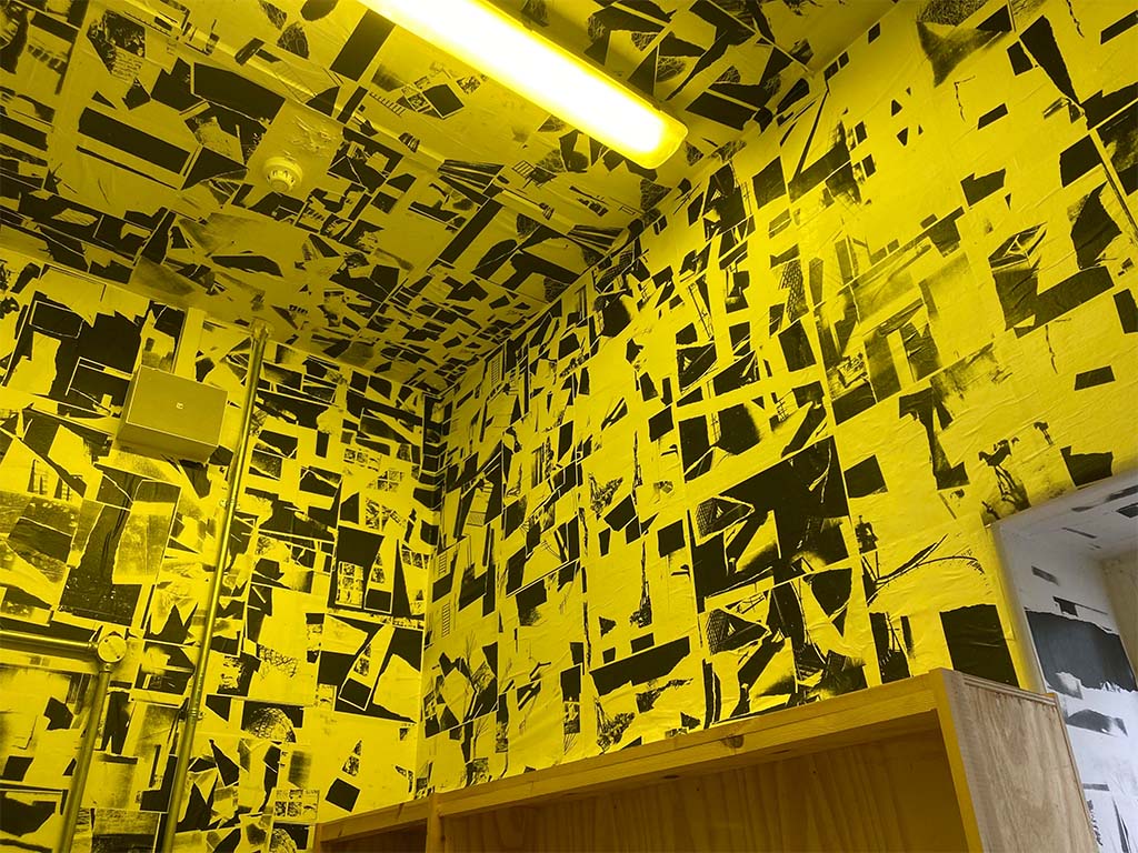 A room in yellow light that is wallpapered in black and white photocopy collages