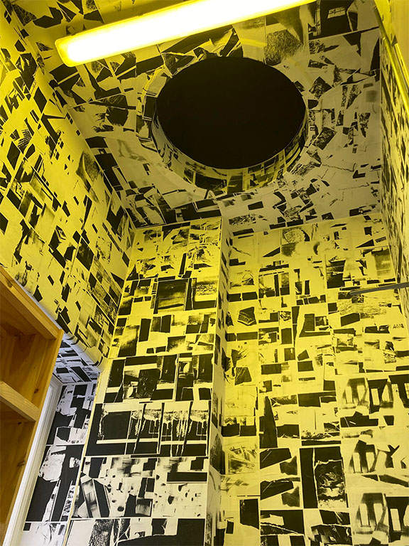 a room lit in yellow light with black and white photocopy collages wallpapered on the walls