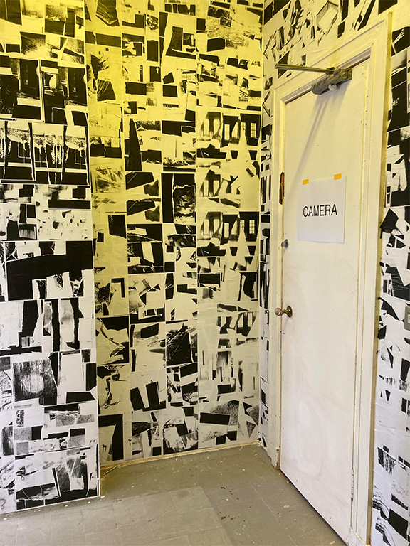 a room lit in yellow light with black and white photocopied collages wallpapered on and a sign that says the word camera taped to a door