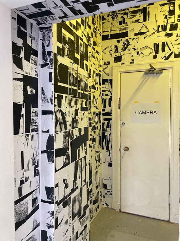 a room lit in yellow light with black and white photocopied collages wallpapered on and a sign that says the word camera taped to a door