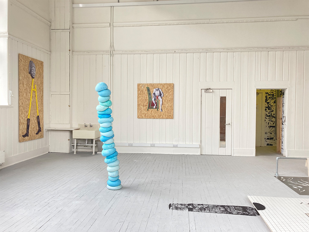 The view of a room featuring various artworks including Two collages made from paper and wood hung on a wall, depicting figures. A sink full of water and pink paint. A blue sculptural column made from jesmonite and an installation with poppy seeds on the floor.