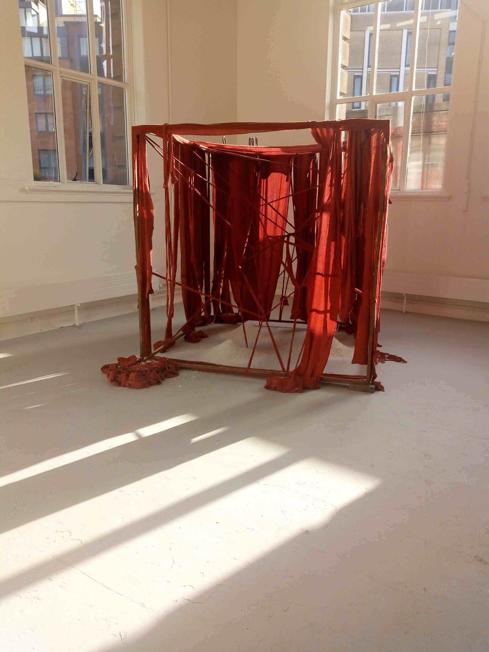 A 2 metre by 2 metre rusted steel frame with internal structure of red fabric criss-crossed and draped and wound with red fabric bandages. There are 3 piles of salt within the structure and 4 upstanding metal hooks at the rear and to the left 