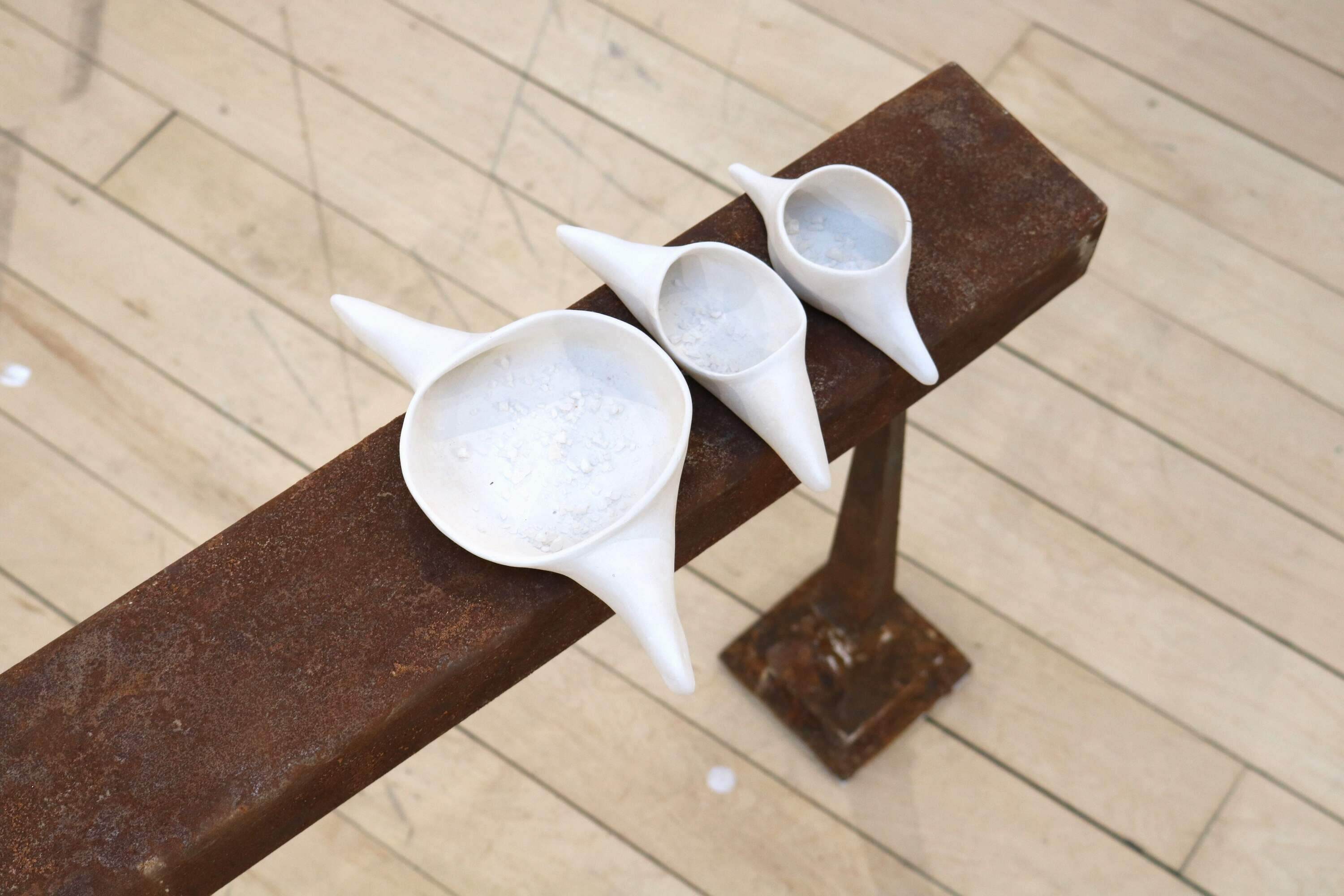A rusted steel narrow bench on narrow legs, 2 metres long, 60 centimetres high and 10 centimetres wide. At one end are placed 3 white ceramic vessels in ascending size containing traces of salt. At the other end of bench there is a sprinkled line of salt. View of ceramic vessels.