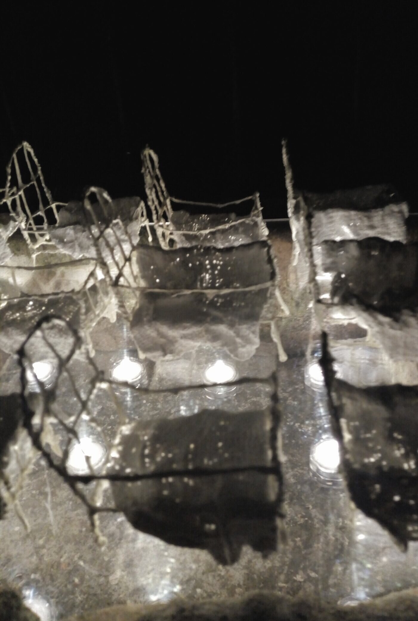 12 tiny wire white model beds with plaster bandage covers in 3 lines of 4 are lit from underneath in the dark above a brick sarcophagus.