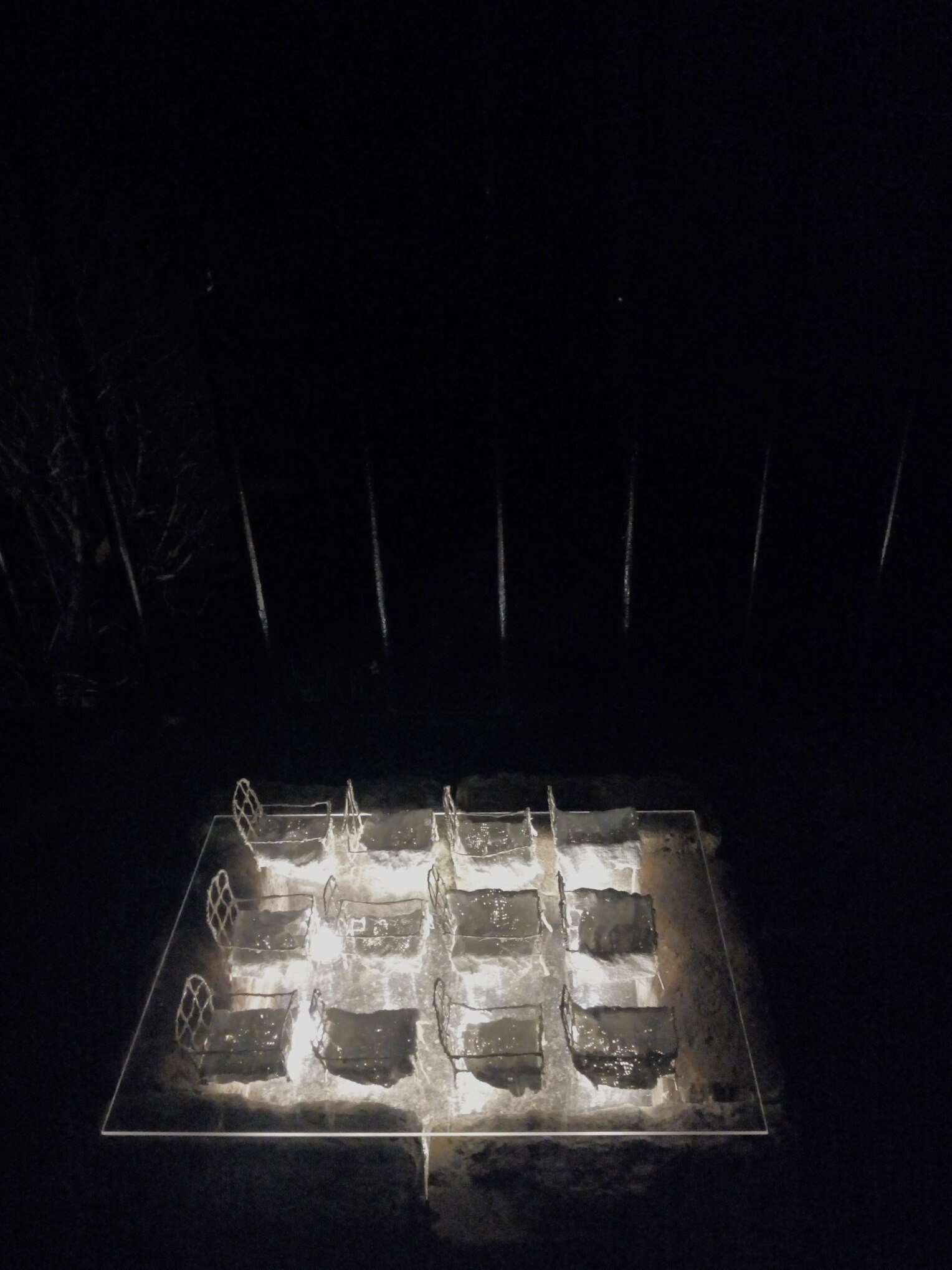12 tiny wire white model beds with plaster bandage covers in 3 lines of 4 are lit from underneath in the dark above a brick sarcophagus. 