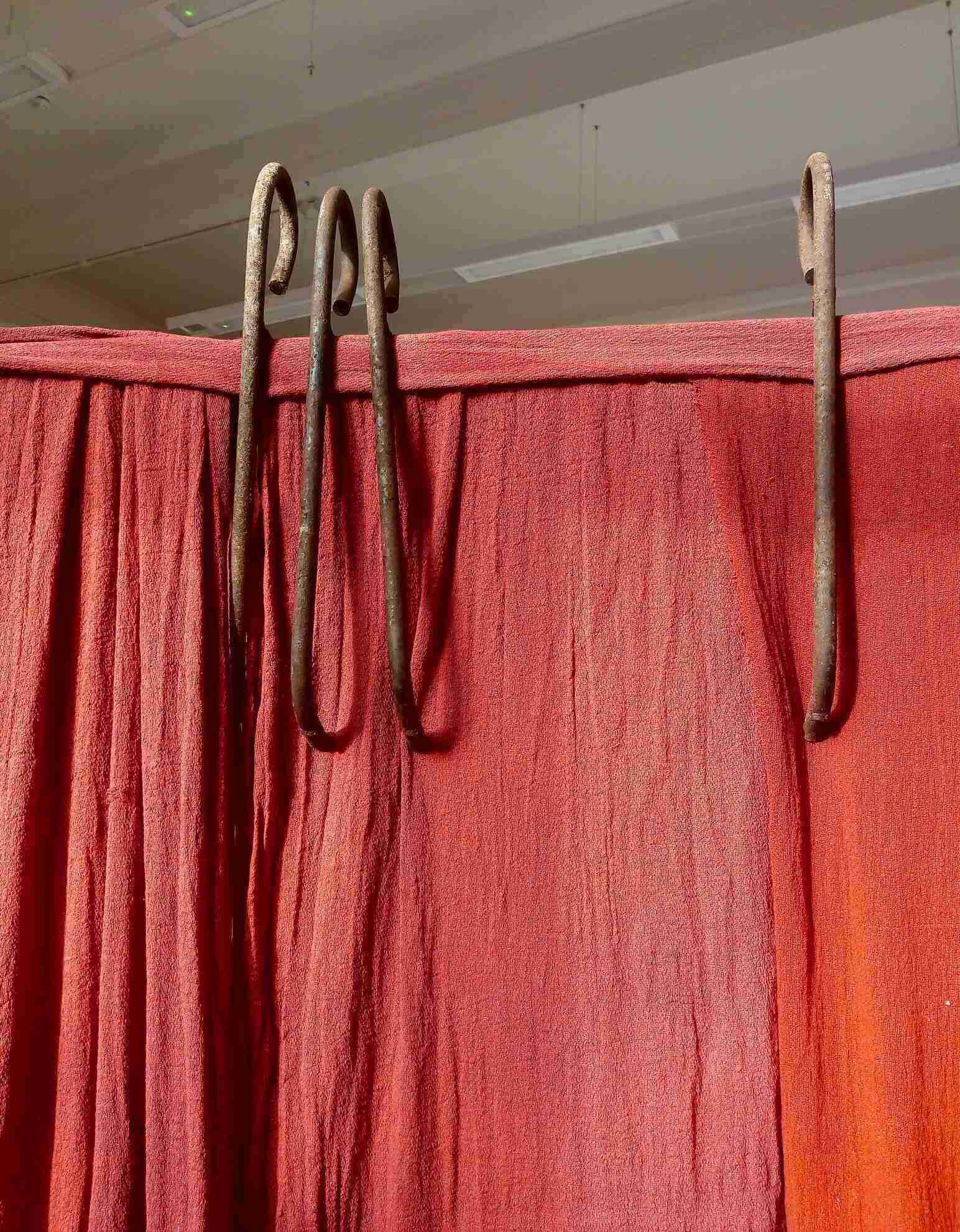 A 2 metre by 2 metre rusted steel frame with internal structure of red fabric criss-crossed and draped and wound with red fabric bandages. There are 3 piles of salt within the structure and 4 upstanding metal hooks at the rear and to the left. Rear view.
