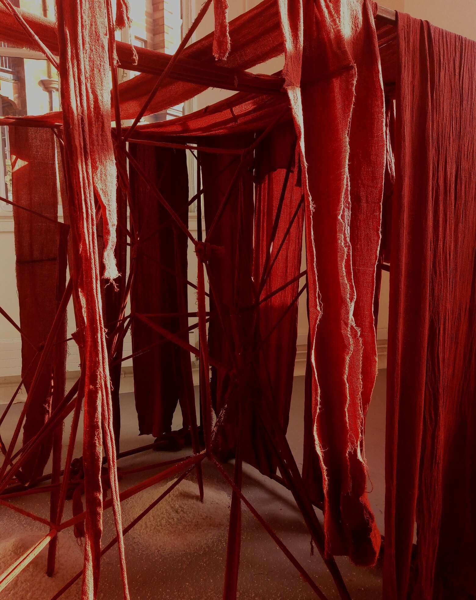 A 2 metre by 2 metre rusted steel frame with internal structure of red fabric criss-crossed and draped and wound with red fabric bandages. There are 3 piles of salt within the structure and 4 upstanding metal hooks at the rear and to the left. Internal view