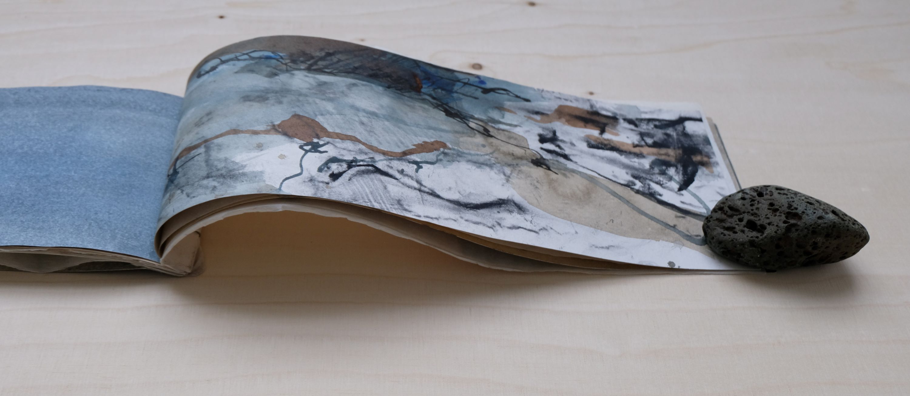 A long handmade book with gestural drawing lays open on a wooden table, a rock holds down the page. 