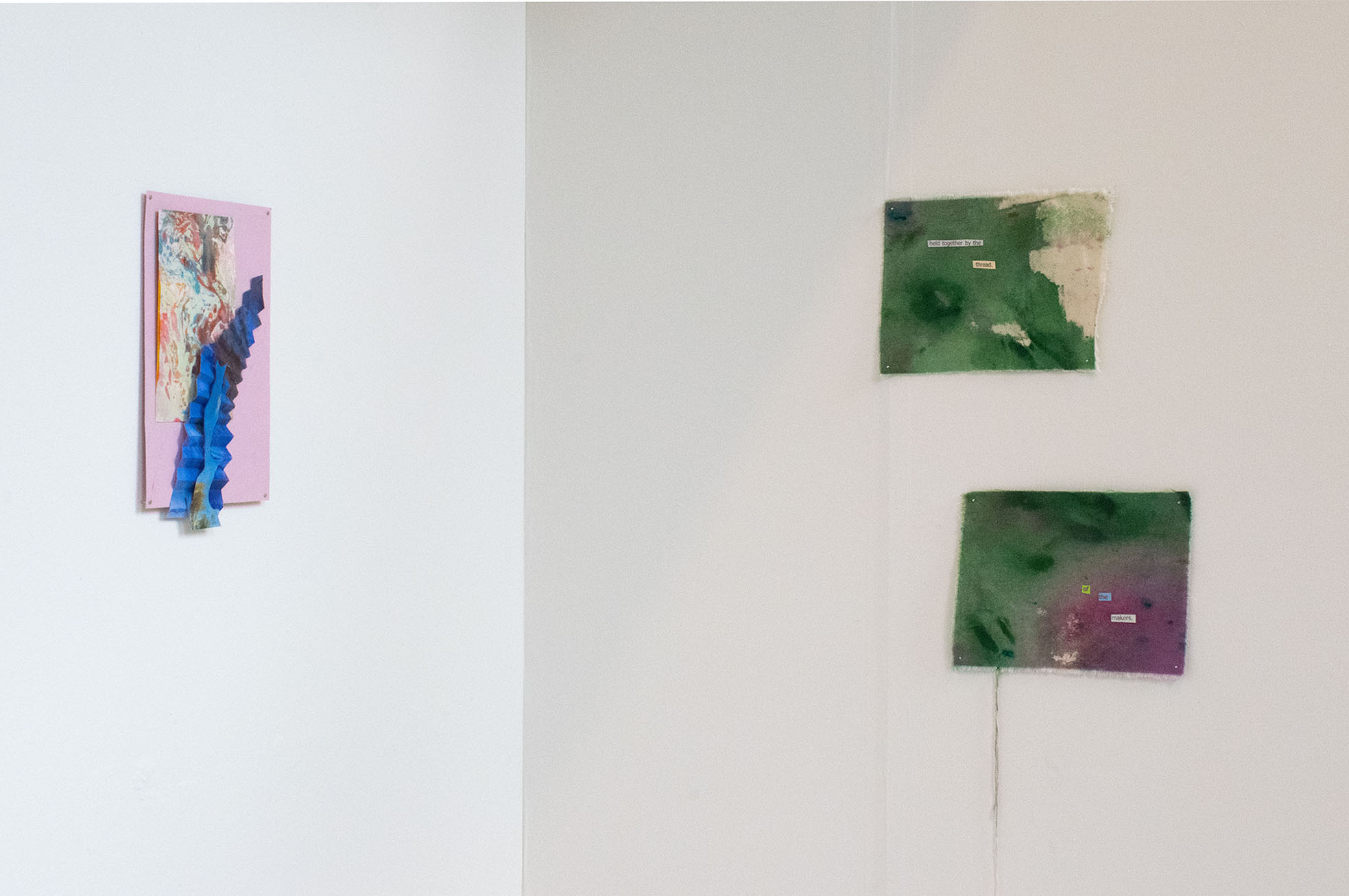 Installation image showing a corner of the space, with a small collage on the left and two abstract green and purple paintings on the right, with collaged text reading "held together by the thread of the makers".