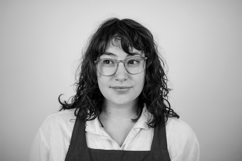 A headshot of Katherine in black and white. She is wearing a white shirt with an apron over the top.