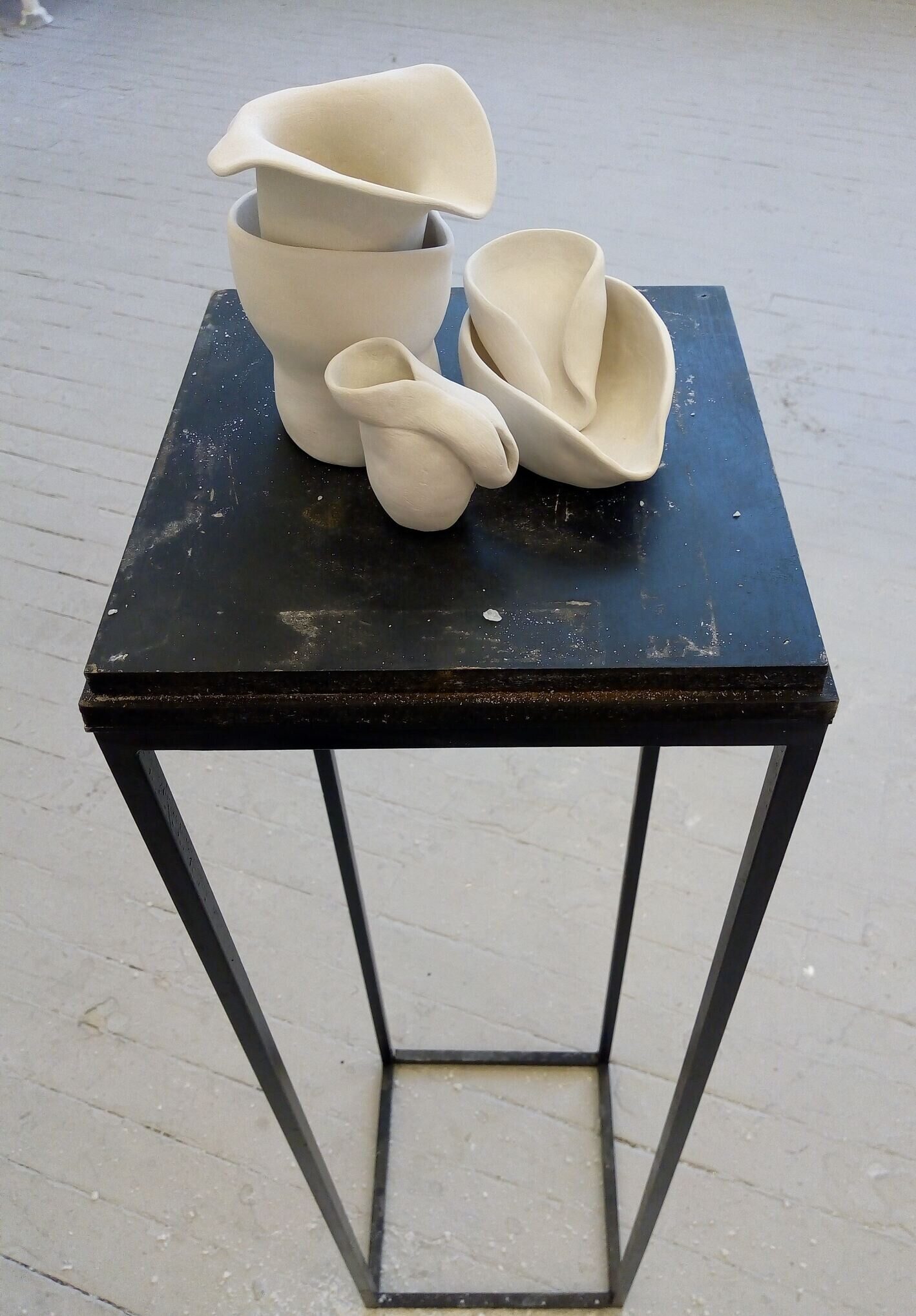 A collection of 5 small white ceramic vessels grouped and piled on one another on a black plinth.