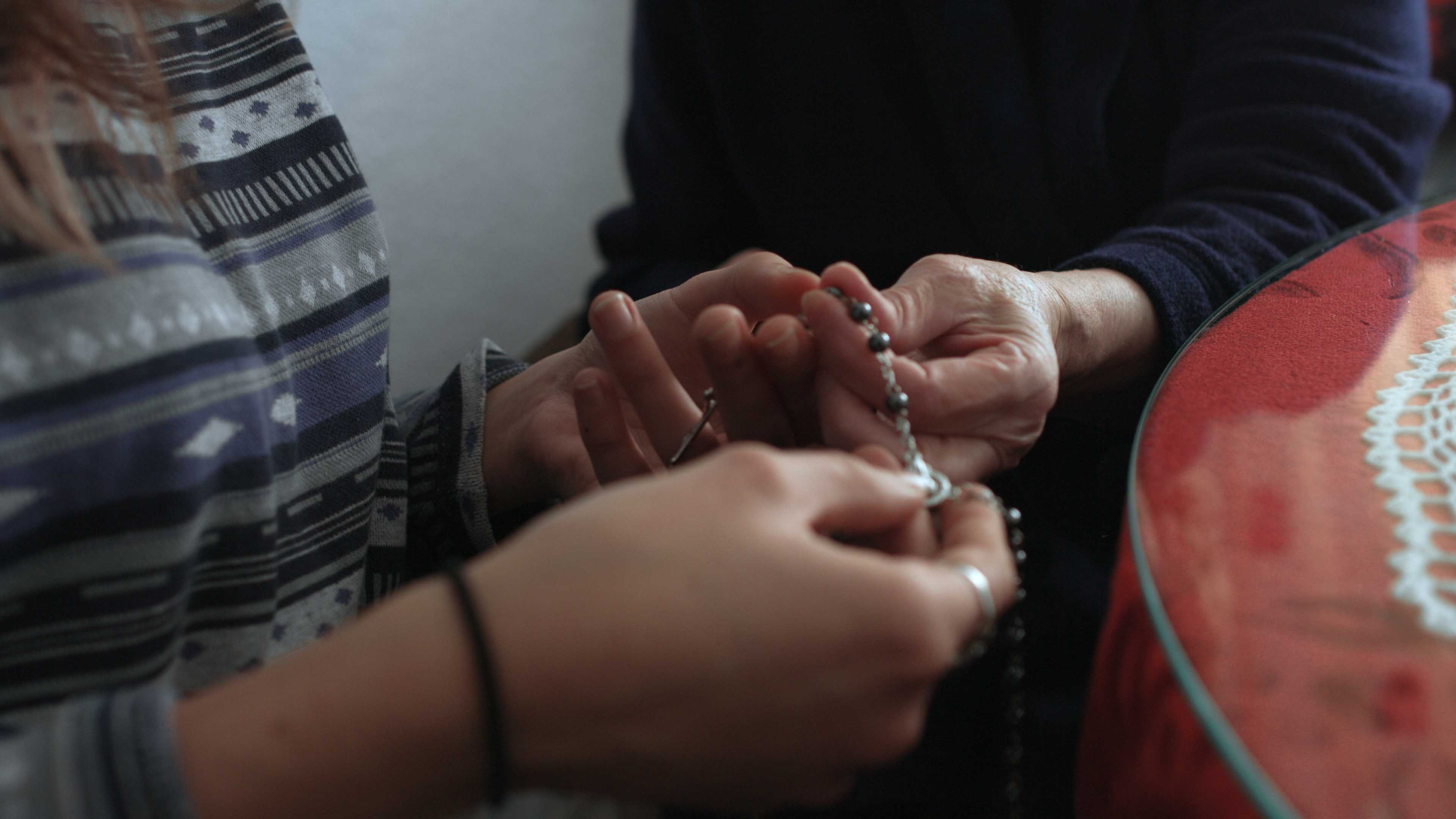 Two pairs of hands holding a rosary, one pair is old, the other young