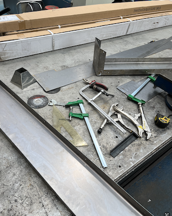 Photo of a work bench in a metal workshop, with clamps and sheets of steel on top of it